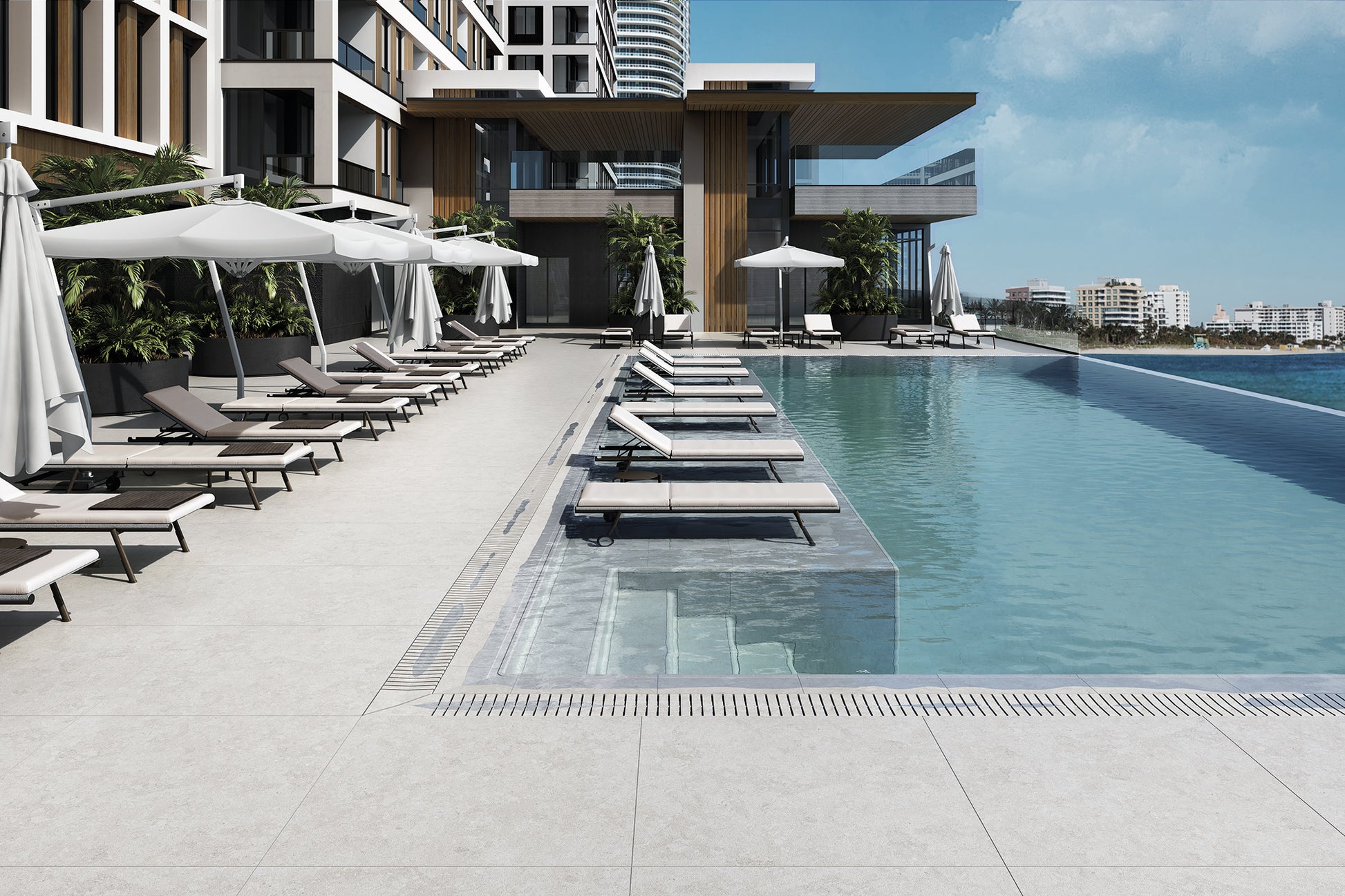 Modern outdoor infinity poolside area with light gray tiled deck, closed white umbrellas, and multiple lounge chairs under a clear blue sky.