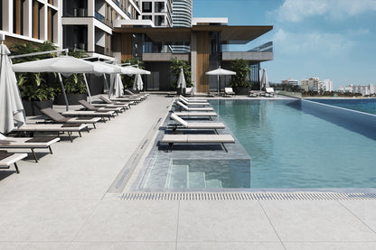 Modern outdoor infinity poolside area with light gray tiled deck, closed white umbrellas, and multiple lounge chairs under a clear blue sky.